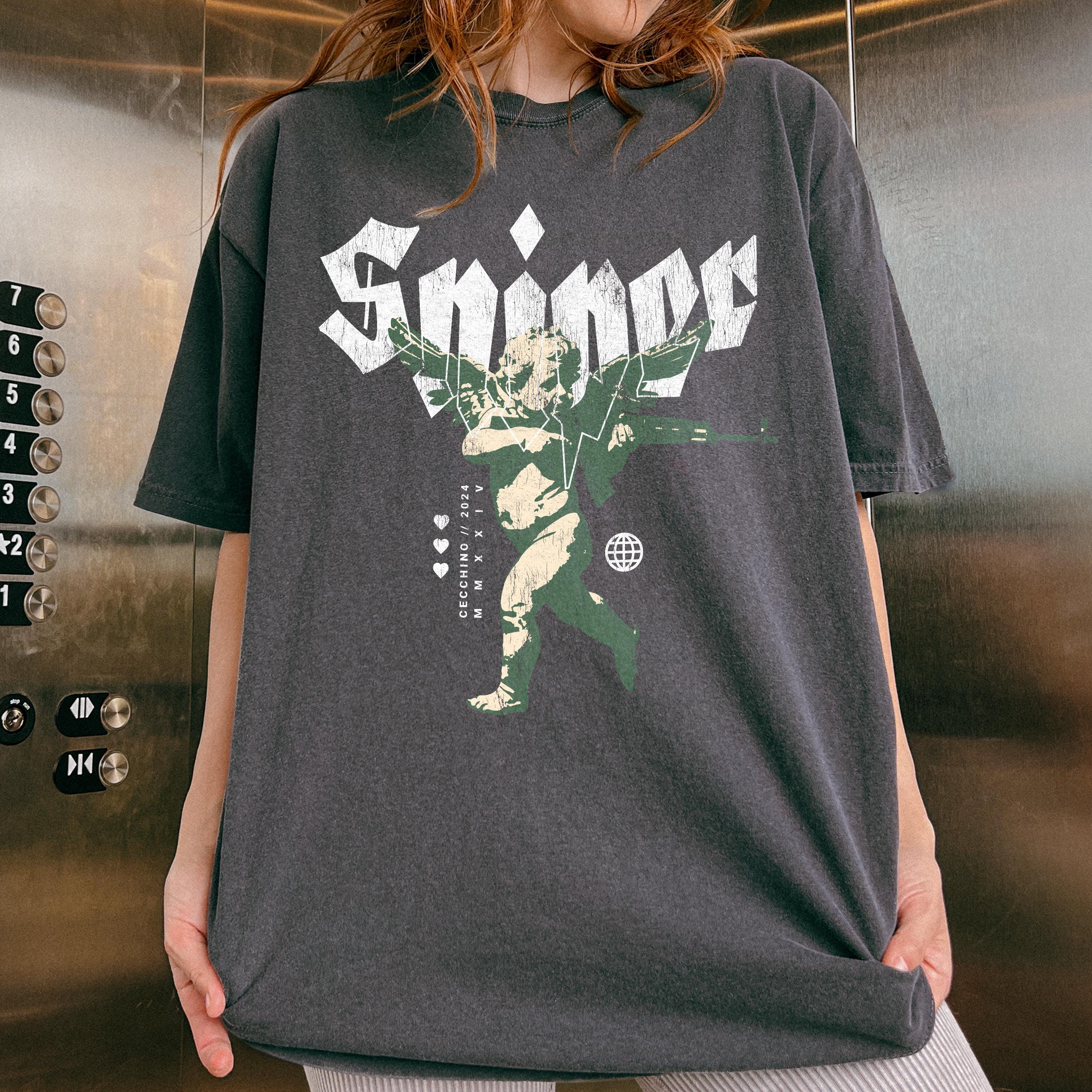 Dark gray oversized t-shirt featuring a graphic design. The design includes the word 'Sniper' in stylized lettering, a cherubic figure wielding a weapon, and small details like hearts and a globe.