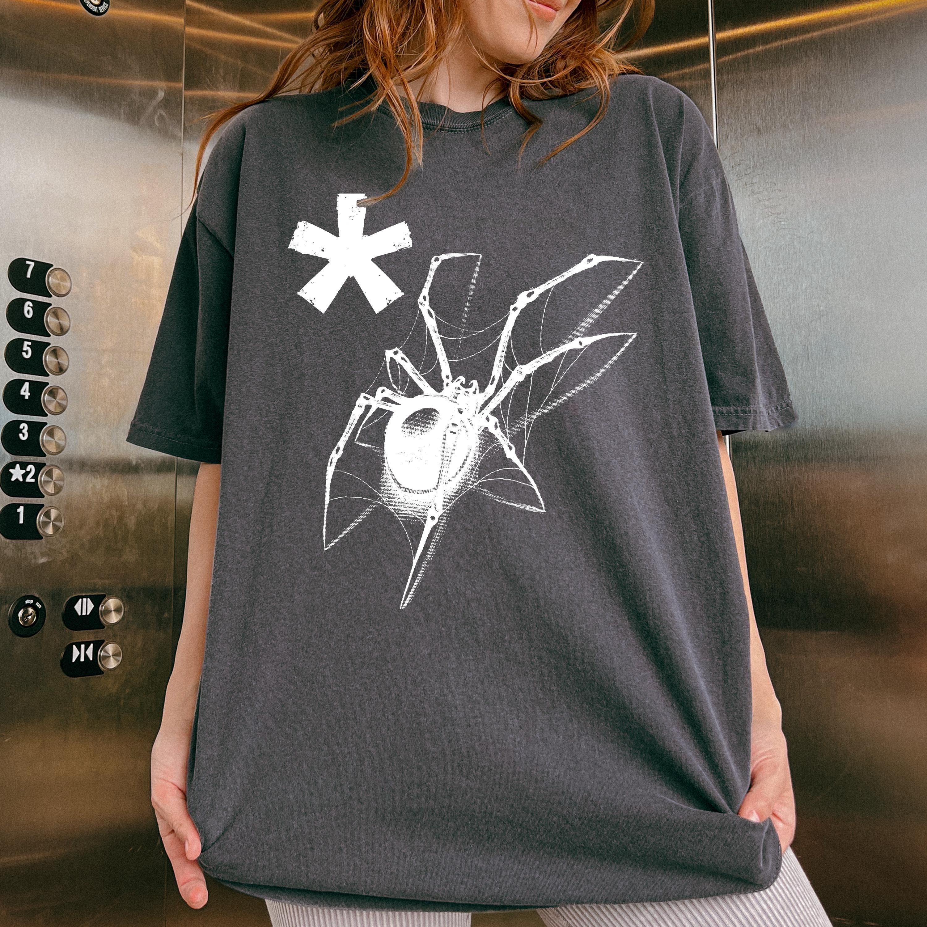 May include: Black oversized t-shirt featuring a graphic design of a spider on a web with a white asterisk. The spider is detailed with thin lines and a white color against the dark shirt. The shirt is made of a soft, comfortable fabric and has a relaxed fit.