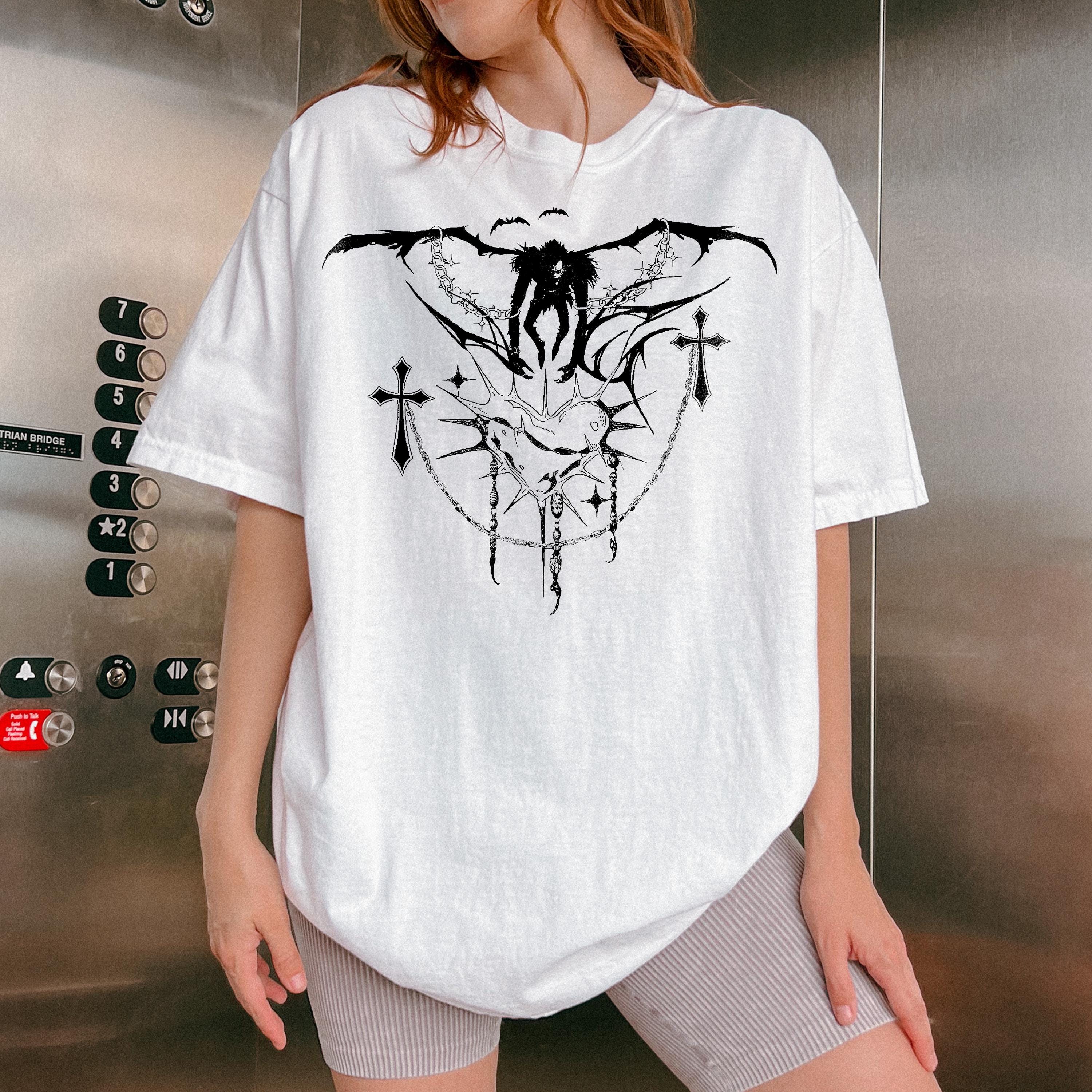 May include: White t-shirt with a black graphic print of a figure with wings and a chain with crosses. The figure is standing on a heart with thorns. The graphic is surrounded by a chain with crosses.