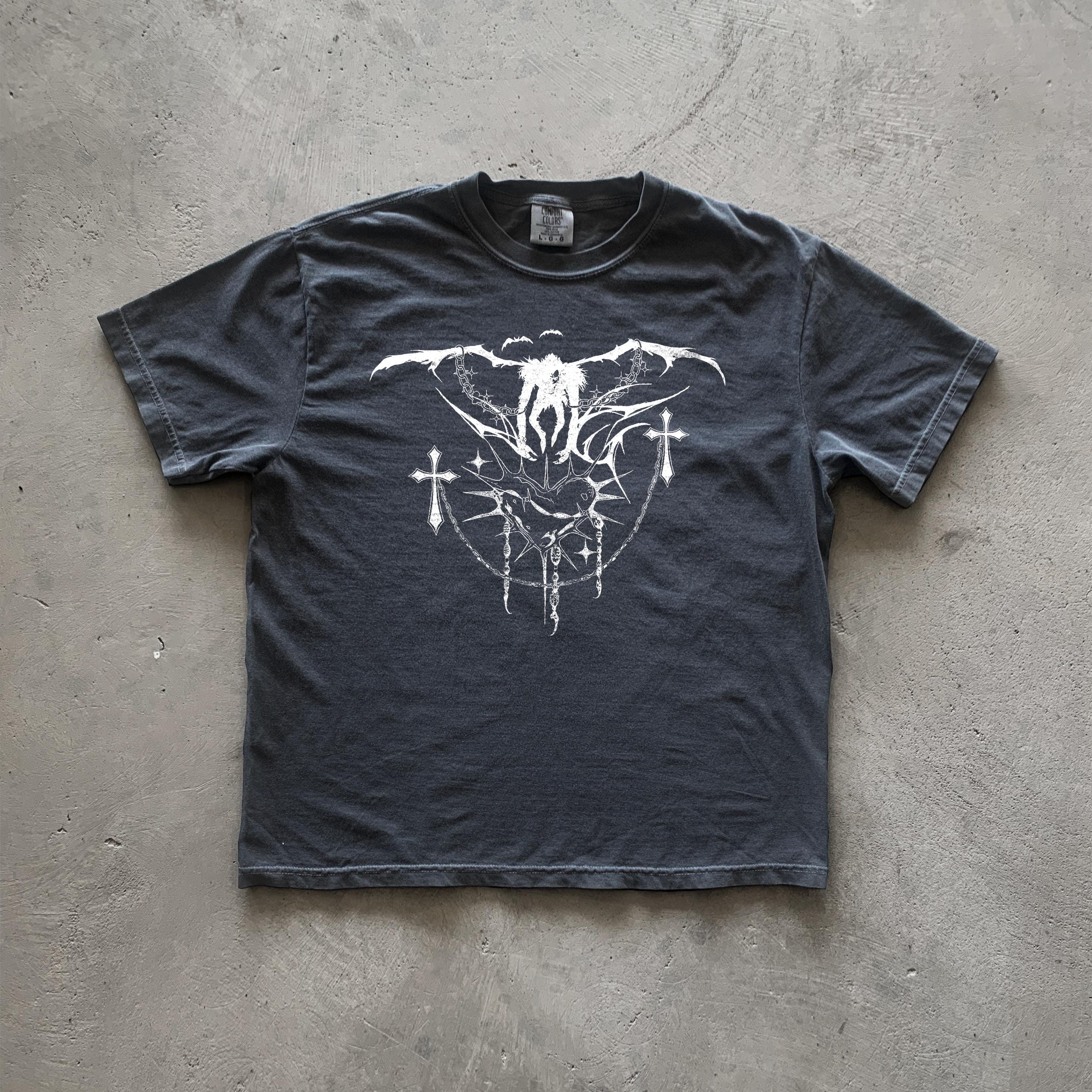 May include: A dark gray t-shirt with a white graphic print of a winged figure with crosses and chains. The graphic is centered on the shirt.